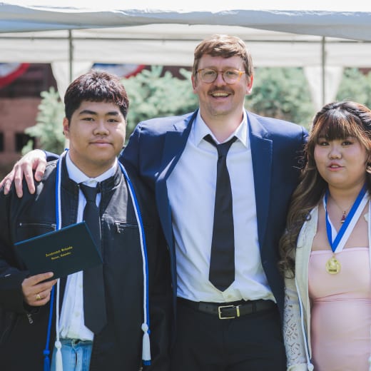 Two graduates holding their diplomas either side of a staff member under the ceremony tent