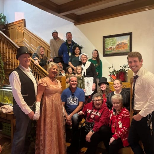 Guests and staff gathered on the mansion staircase, several in Victorian costume, beside the decorated Christmas tree
