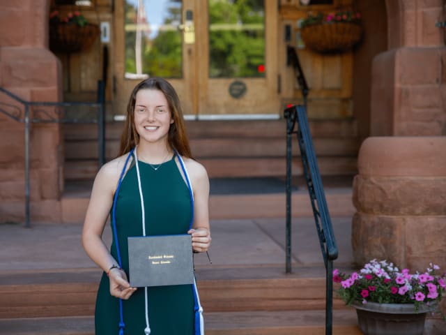 A graduate in a teal dress holding their diploma beside the flowers on the mansion steps