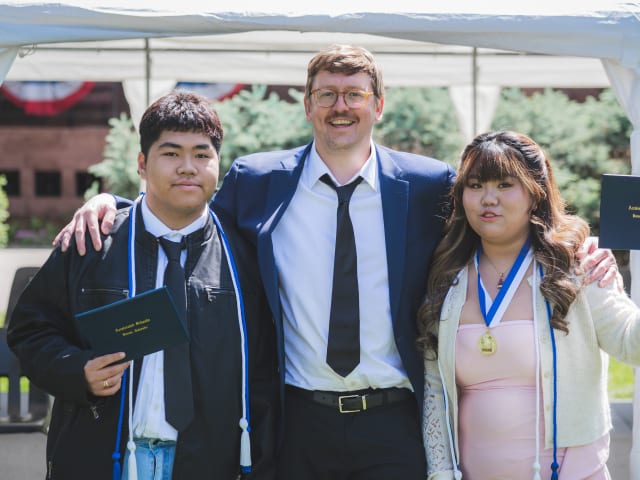 Two graduates holding their diplomas either side of a staff member under the ceremony tent