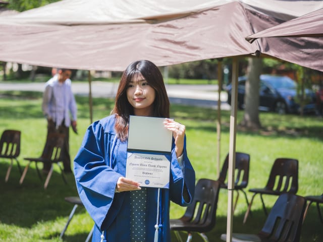 A graduate in a blue gown holding up her Accelerated Schools diploma on the lawn