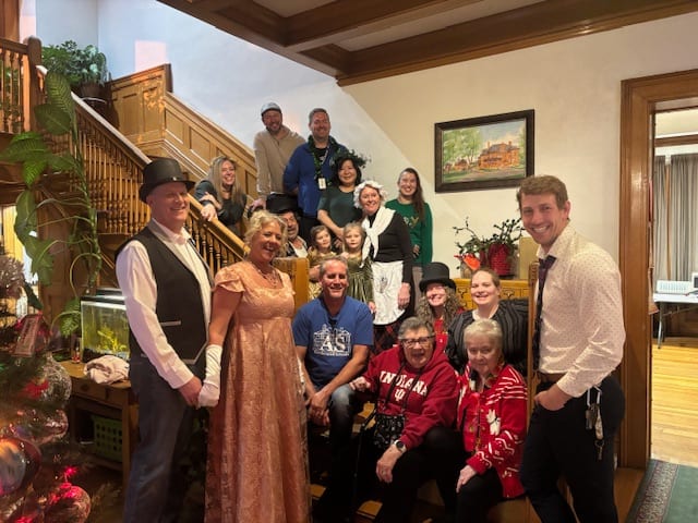 Guests and staff gathered on the mansion staircase, several in Victorian costume, beside the decorated Christmas tree