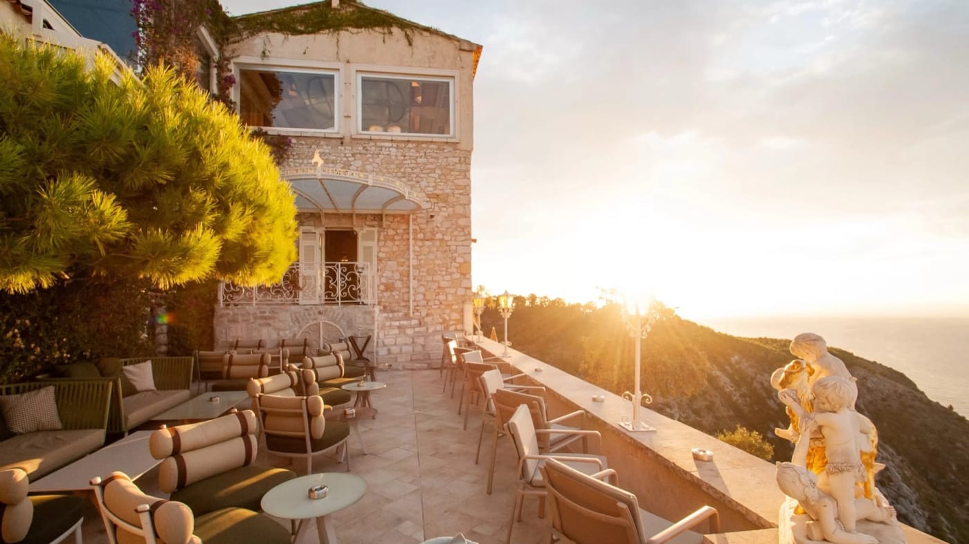 Bar terrace of the Château de la Chèvre d'Or in Èze at sunset, armchairs, statue and the sea below