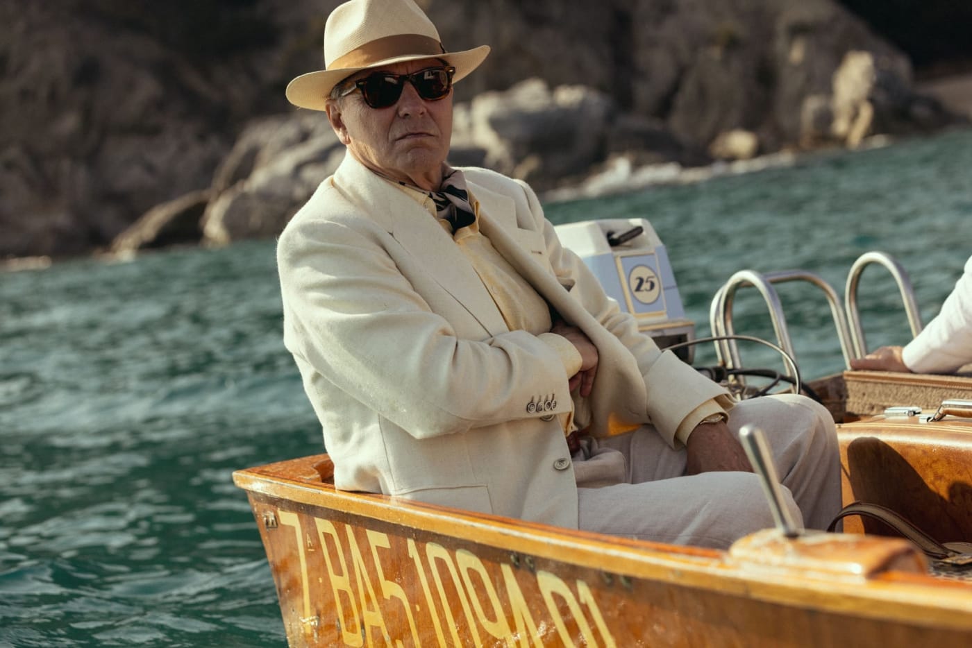 José Coronado de fato creme e chapéu, sentado num barco, em El problema final