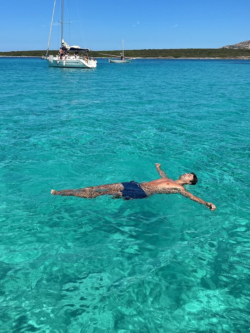 Raphaël Simon flottant dans une eau turquoise, des voiliers au mouillage à l'arrière-plan