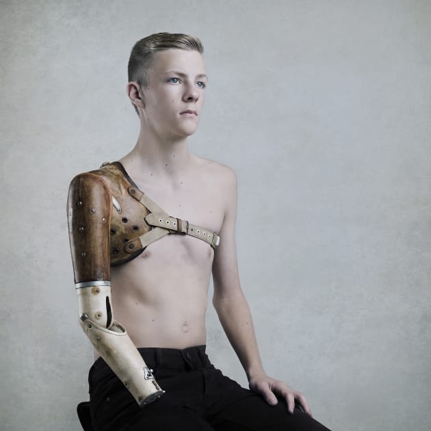 Cameron and the prosthetic arm, 2016 by Steve Wise.
This image is part of a series of portraits exploring the collection of historical medical equipment and artefacts housed in the Royal Perth Hospital Museum. Cameron is able-bodied, with the full use of both arms. He is naturally right-handed, and is a very successful young sportsman, so when I asked him to wear the prosthetic I was anticipating an interesting result. On suggesting he imagine not having the use of his right arm, his face took on an expression that was hard to read – perhaps the envisioning of change, loss. This added a unique mood to his character in this formal portrait.