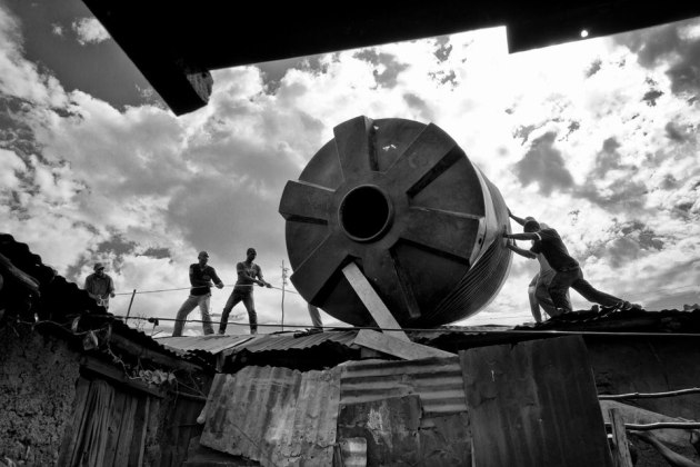 © Maureen Ruddy Burkhart. Moving the 10,000 Litre Water Tank, Kenya. The Kibera slum outside of Nairobi has no running water or plumbing for between 500,000 and 1 million residents. Toilets are limited to a few key areas served by these huge tanks. To get this tank to its destination many men were hired to roll the (empty) tank over flimsy metal rooftops.