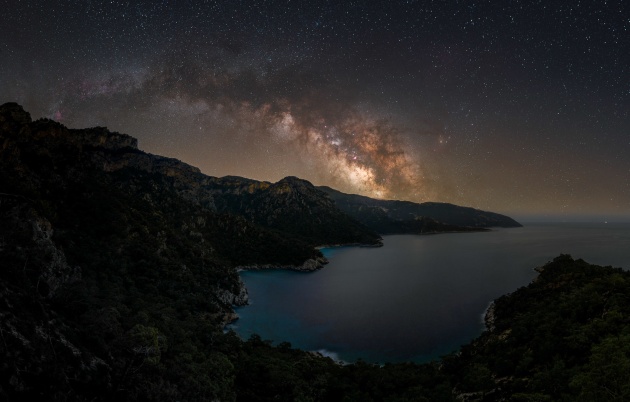 © Alyn Wallace - “Paradise Beach” 
Lycian Way, Turkey
I spent the pandemic in a remote valley on the southwest coast of Turkey trying to avoid the chaos of restrictions and lockdowns and be surrounded by beautiful nature instead. Sadly, the valley I was residing in has very steep walls and only a clear view west, but, as the Milky Way core rises in the southeast at this time of year, I had to hike up over the mountains to get a good view.
The path to get to this location is part of the Lycian Way, a popular multi-day hiking trail along the incredibly beautiful coastline of Turkey. The view overlooks a beach known as “Cennet,” which, translated from Turkish, means “Paradise,” a name I completely agree with!
Sitting on the mountainside enjoying this view of the Milky Way arching across the mountains whilst listening to the sound of the waves breaking gently below and the owls twitting through the valleys was just the kind of peace and solitude I needed.