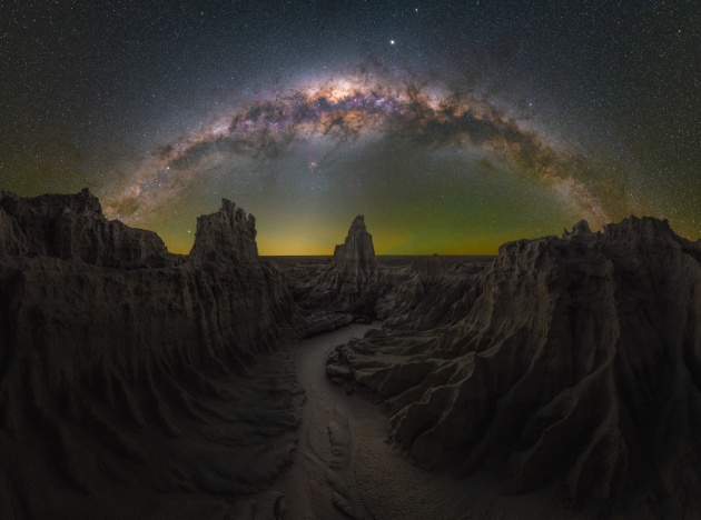 © Daniel Thomas Gum - “Dragon’s Lair” 
Mungo, NSW – Australia
This is my favourite nightscape image to date. Mungo is a 12-hour drive away from my home in Sydney, but those Bortle 1 skies are the best I’ve ever witnessed and photographed at night. I had perfect conditions for three straight nights, with really good seeing throughout.
The moment I came upon this scene, I knew exactly what I wanted to the name the image. It was otherworldly – think Game of Thrones – and it lined up perfectly for how I wanted to capture it. Large, jagged walls framed a winding path leading to a centered spire to the west. There was only ever going to be one way to do it justice and that was as a multi- layered Milky Way panorama.
I planned this image using PhotoPills during the day, but in post-processing, I decided to use the blue-hour blend for the foreground with a tracked sky for the cleanest possible image.