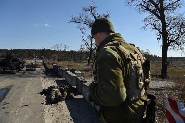 © Kate Geraghty. Winner, Feature/Photographic Essay. Invasion of Ukraine - Civilian Impact. Frontline aftermath: On a frontline position hundreds of metres from Russian forces, a Ukrainian soldier with the 72nd Brigade stands guard over the bodies of two Russian soldiers that have been recovered from a battleground. The Russian soldiers’ bodies are two of six that will be handed over to the Red Cross in an exchange for two Ukrainian soldier prisoners of war. Not long after this photo was taken. Russian Grad missiles hit this position, killing three Ukrainian soldiers. Kyiv Oblast, Ukraine. March 18, 2022.