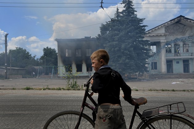 © Kate Geraghty. Winner, Feature/Photographic Essay. Invasion of Ukraine - Civilian Impact. Lysychansk: As the sound of one of four incoming missiles whistles ahead, eight-year-old Timosha stands with his bicycle in front of the still burning Palace of Culture in Lysychansk. The previous day, an air-strike hit the Palace, killing four people. At the time this photo was taken Lysychansk was the last Ukrainian-held city in the Luhansk Region; a week later the city and its remaining residents were under Russian occupation. Lysychansk, Ukraine. June 17, 2022.