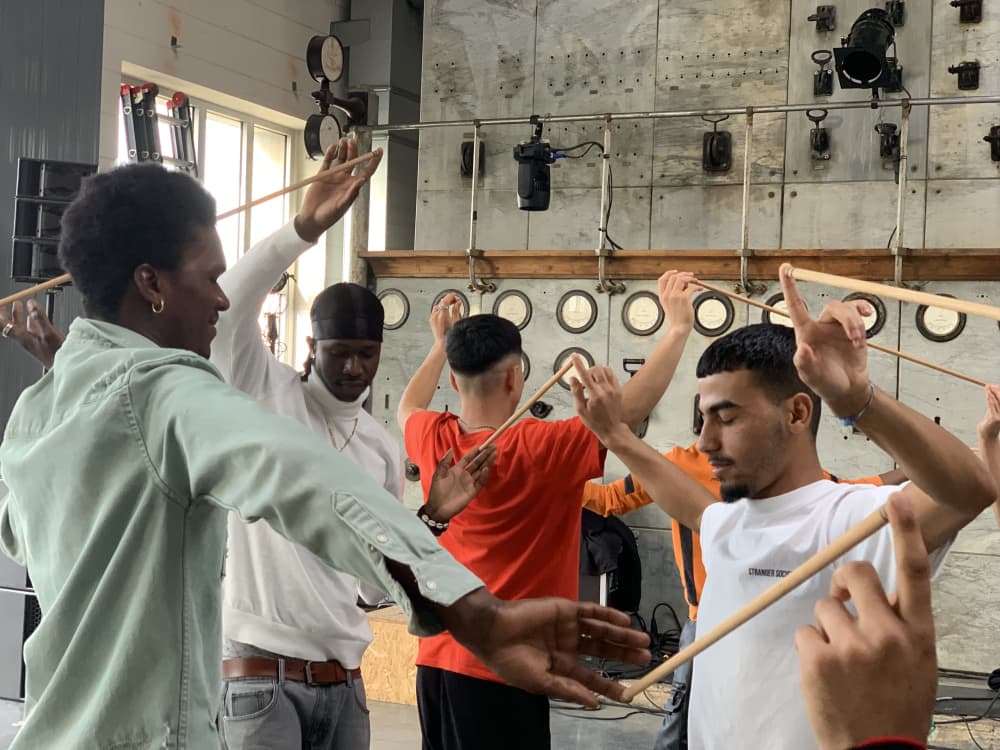 A group of young people, eyes closed, collaborates on a game-like task by holding sticks pressed between their hands.