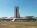 Brazil's National Congress in Brasília under a clear blue sky, with the twin office towers between the Senate dome and the Chamber of Deputies bowl, behind a wide dry lawn