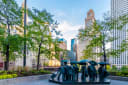 Bronze sculpture of figures holding umbrellas on a plaza in downtown Chicago, with trees and office towers behind under a blue sky