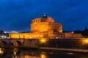 Castel Sant'Angelo in Rome lit up at dusk above the River Tiber, with the Ponte Sant'Angelo to the left under a deep blue sky
