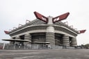 The San Siro stadium in Milan, with its red roof girders and spiral ramp towers, under a pale overcast sky