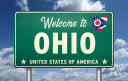 A green Welcome to Ohio road sign with the Ohio flag emblem and the words United States of America, set against a blue sky with clouds