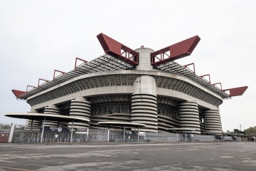 The San Siro stadium in Milan, with its red roof girders and spiral ramp towers, under a pale overcast sky