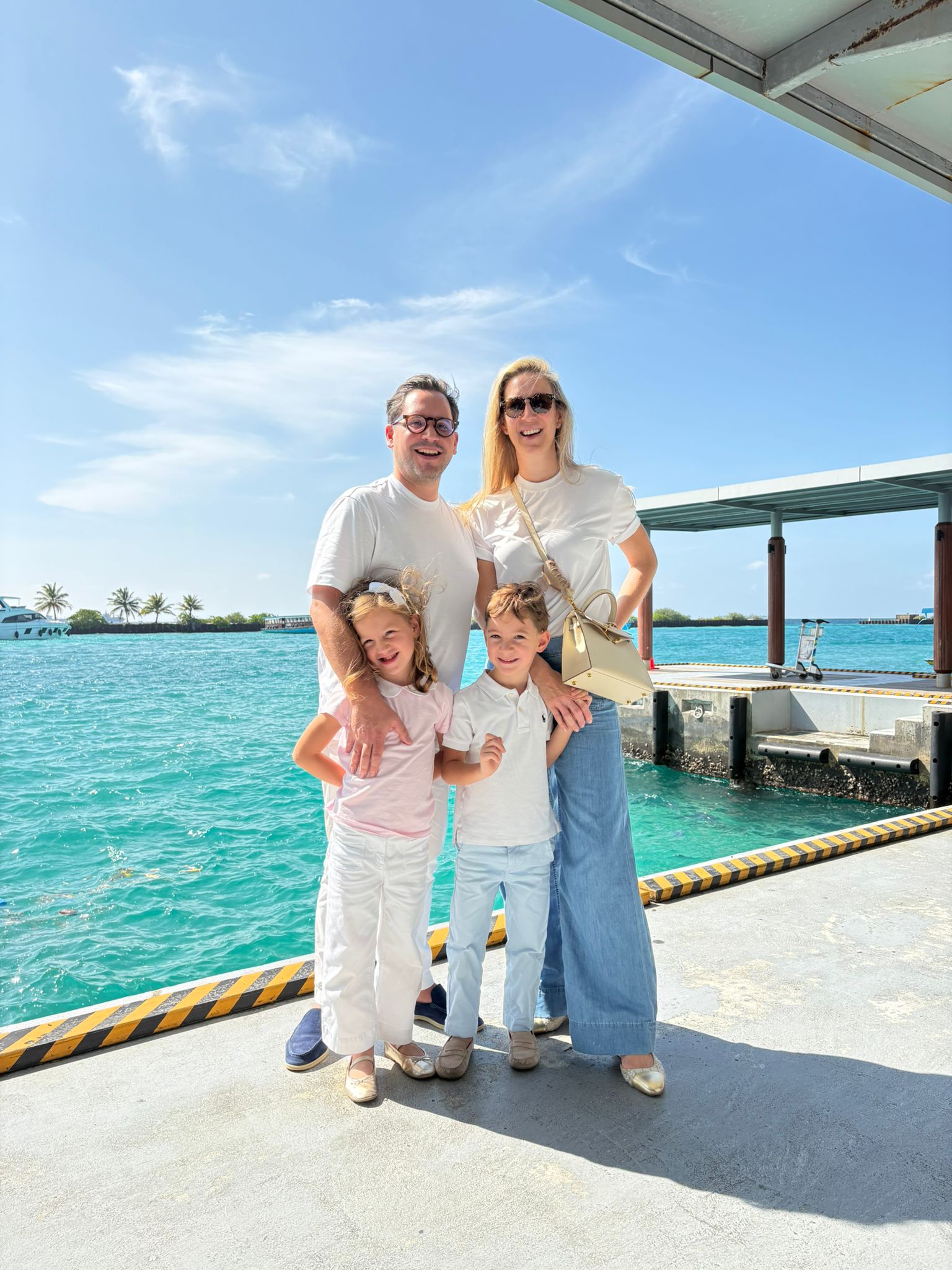 A family of four stands smiling on a concrete pier next to bright ocean water, with a distant island and boats visible under a sunny blue sky.