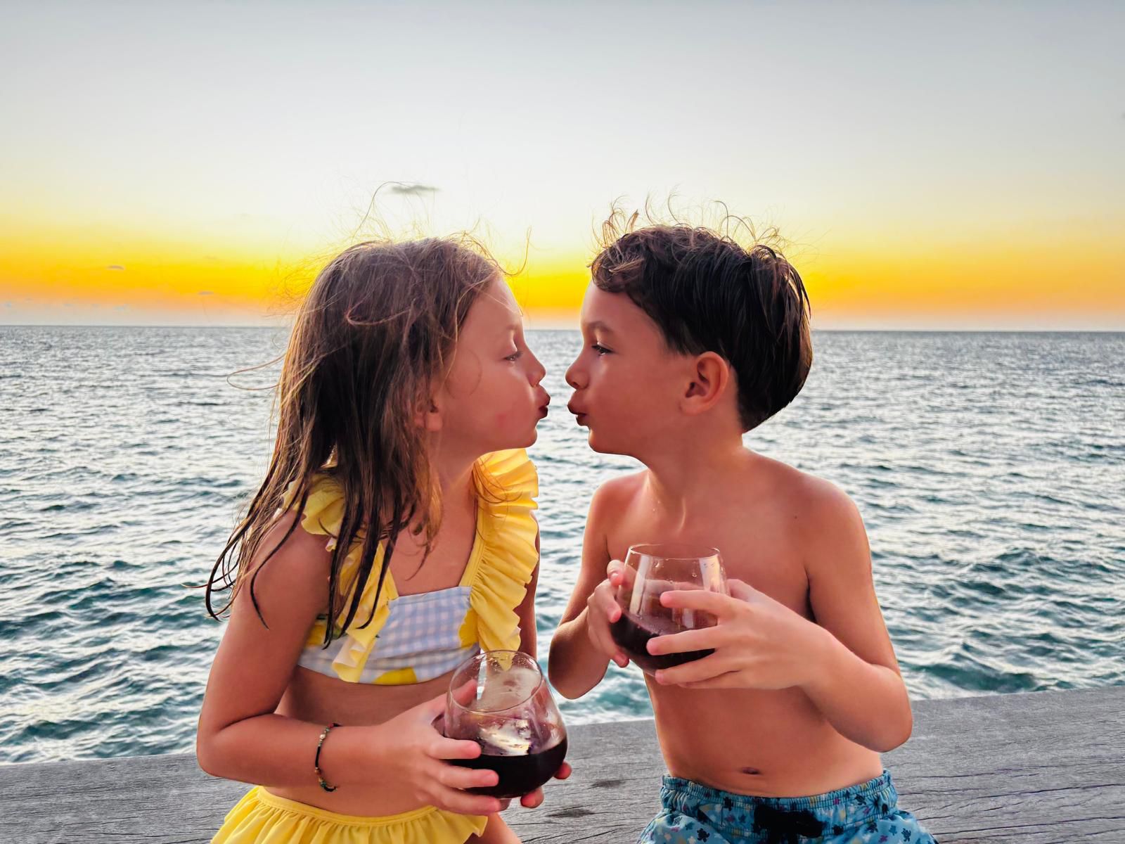 A young girl in a yellow swimsuit and a boy in blue trunks sit on a wooden deck by the ocean at sunset, holding dark drinks and leaning in for a kiss.