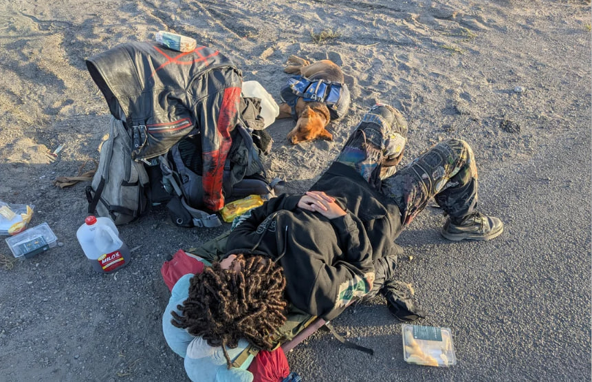 A person with dreadlocks lies on their back on a gravel roadside, resting with hands on stomach, surrounded by a large backpack, leather jacket, gallon jug of water, food containers, and scattered gear. A small brown dog in a blue vest lies curled up nearby, asleep in the dirt.