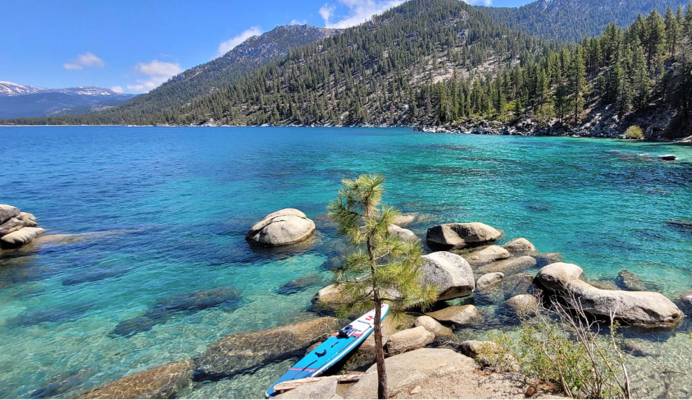 A stunning view of crystal-clear turquoise water in a mountain lake, with large smooth granite boulders and a small pine tree in the foreground. A blue inflatable paddleboard leans against the tree on the rocky shore. In the background, dense evergreen forest climbs steep mountains under a bright blue sky.