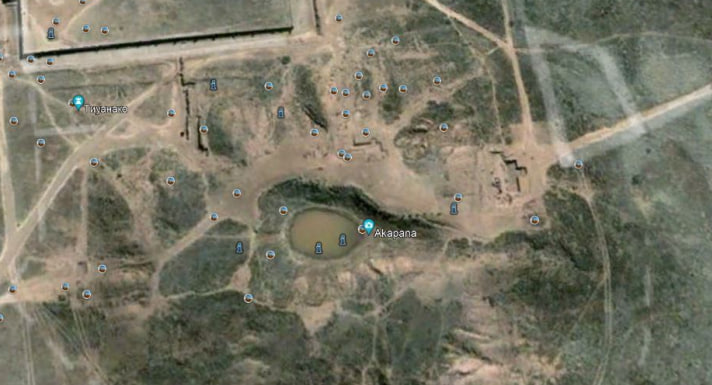 Satellite aerial view of the central Tiwanaku archaeological site in Bolivia, showing the Akapana pyramid mound in the center with a small pond or crater at its top, the rectangular Kalasasaya complex to the northwest, Putuni to the west, and the broader excavated area with scattered ruins and paths in the arid high-altitude landscape.