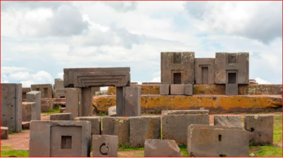 Scattered massive precision-cut andesite stone blocks at Puma Punku in the Tiwanaku complex, Bolivia, including distinctive H-shaped clamps, recessed grooves, and partially reconstructed gate-like structures with T-shaped and rectangular niches, set against a cloudy sky in the high-altitude plain.