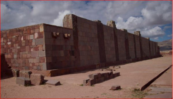 Long, precisely constructed wall at Puma Punku (Pumapunku) in the Tiwanaku archaeological complex, Bolivia, made of large reddish andesite blocks with vertical pilasters, protruding tenon stone heads near the top, scattered stone blocks in the foreground, under a cloudy sky in the barren Altiplano landscape.