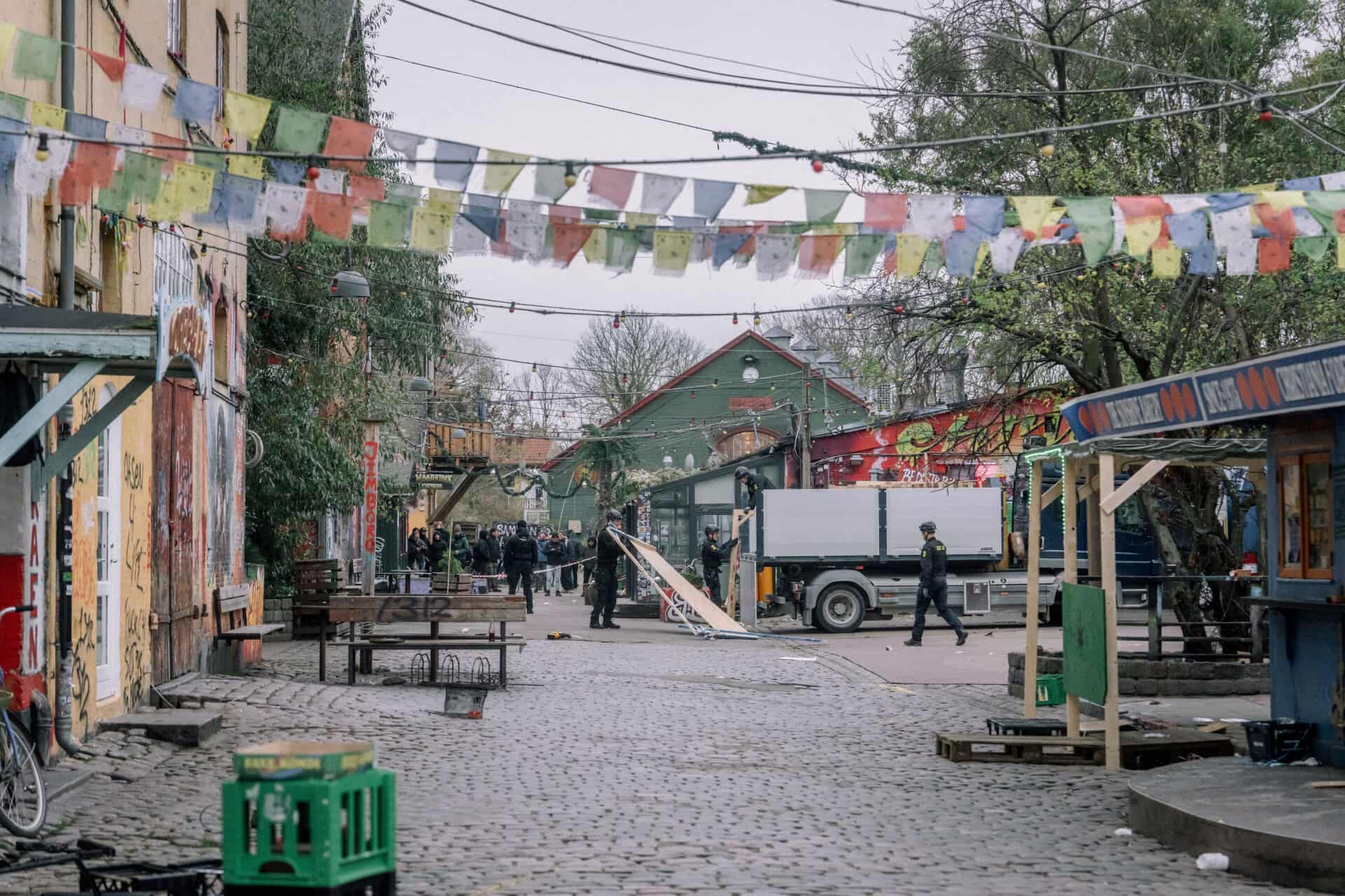 Street scene in Christiania, Copenhagen with colorful prayer flags, graffiti-covered buildings, cobblestone road, and police presence in the background