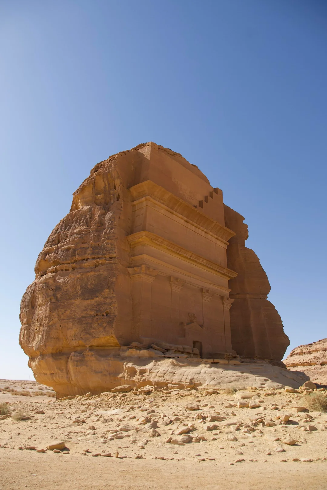 A large sandstone rock with an ancient carved façade standing alone in a desert under a clear blue sky.