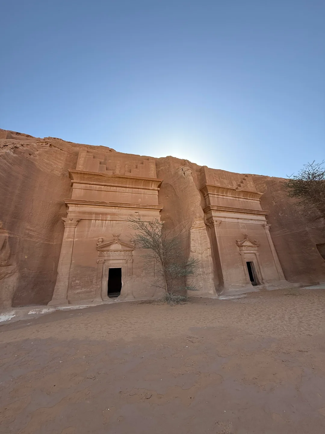 Ancient rock-cut tombs carved into a sandstone cliff under a clear blue sky, with a small tree in front.