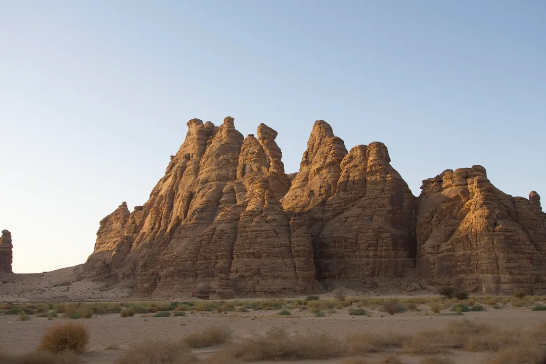 Dramatic cluster of towering, eroded sandstone rock formations with jagged peaks rising from a sandy desert floor under a clear blue sky in AlUla, Saudi Arabia.