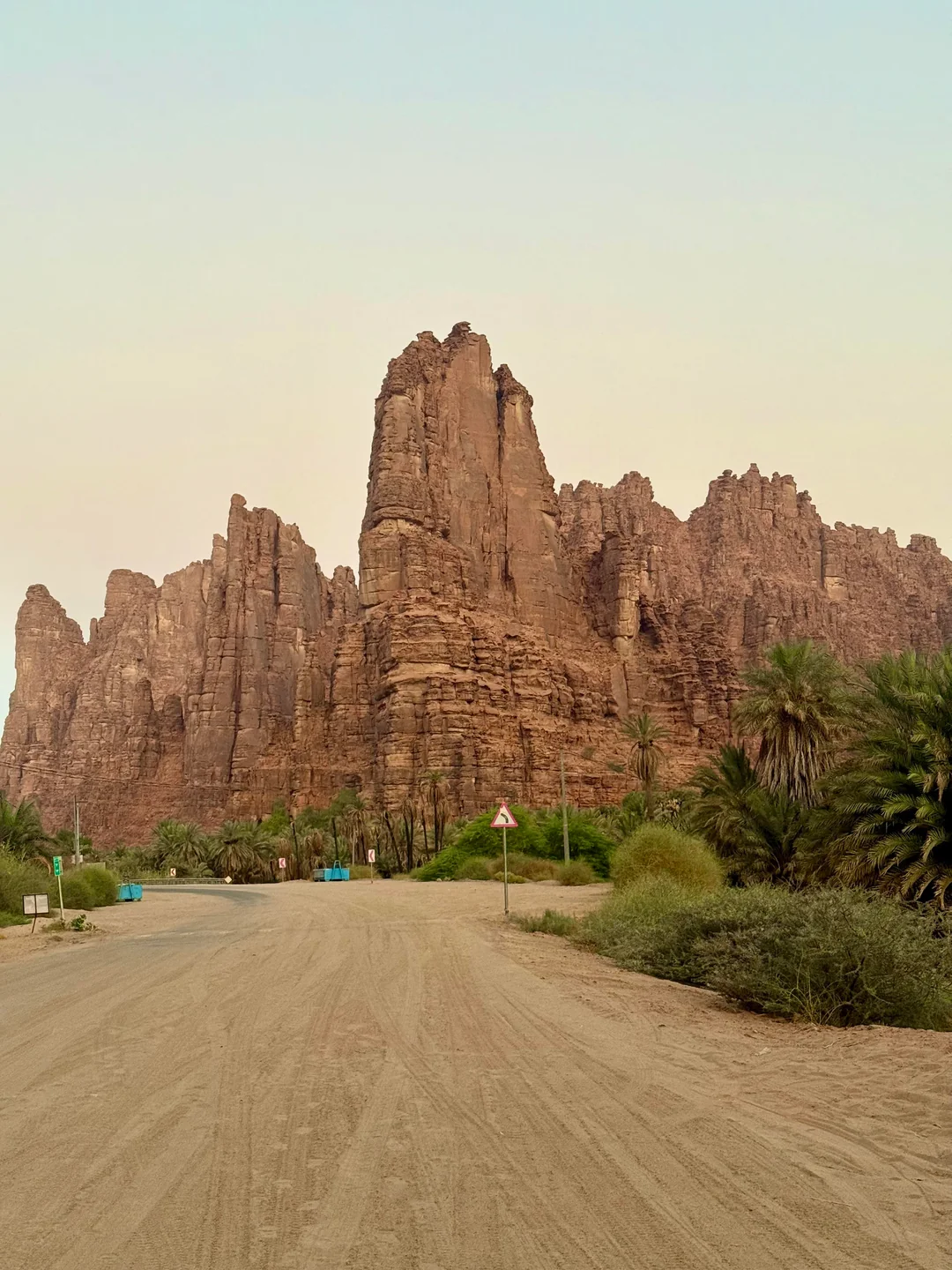 A towering, jagged sandstone cliff rises dramatically above a palm-filled oasis, with a dirt road and yield sign in the foreground, set against a hazy desert sky in AlUla, Saudi Arabia.