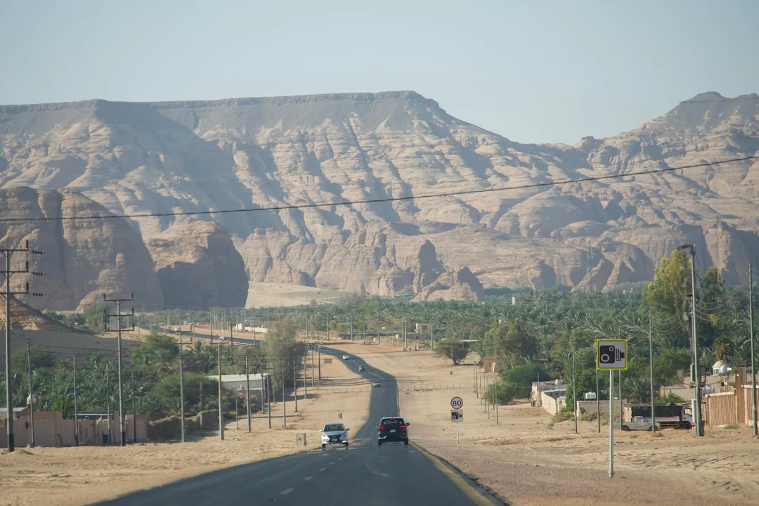 A straight desert highway flanked by palm trees and low buildings, leading toward dramatic layered sandstone mountains under a clear sky in AlUla, Saudi Arabia.
