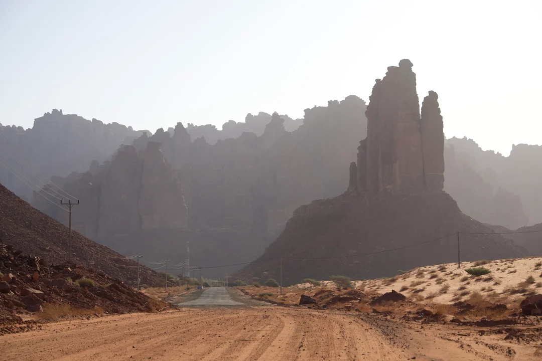 A desert road stretches toward dramatic sandstone cliffs and towering rock pillars under a hazy sky, with power lines running parallel to the track in a remote arid landscape, likely in the Hisma Basin or near Wadi Rum, Jordan.