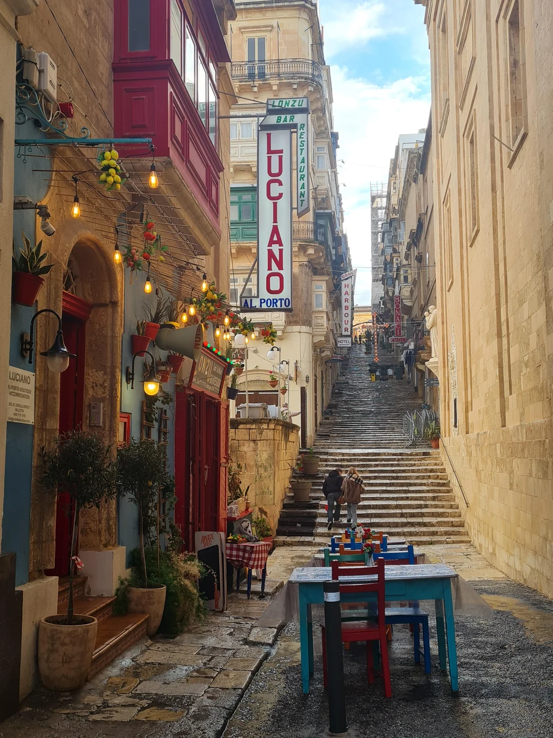 "Charming narrow street in Malta with outdoor café seating and a steep stone staircase ascending between historic buildings, featuring colorful balconies and restaurant signs."