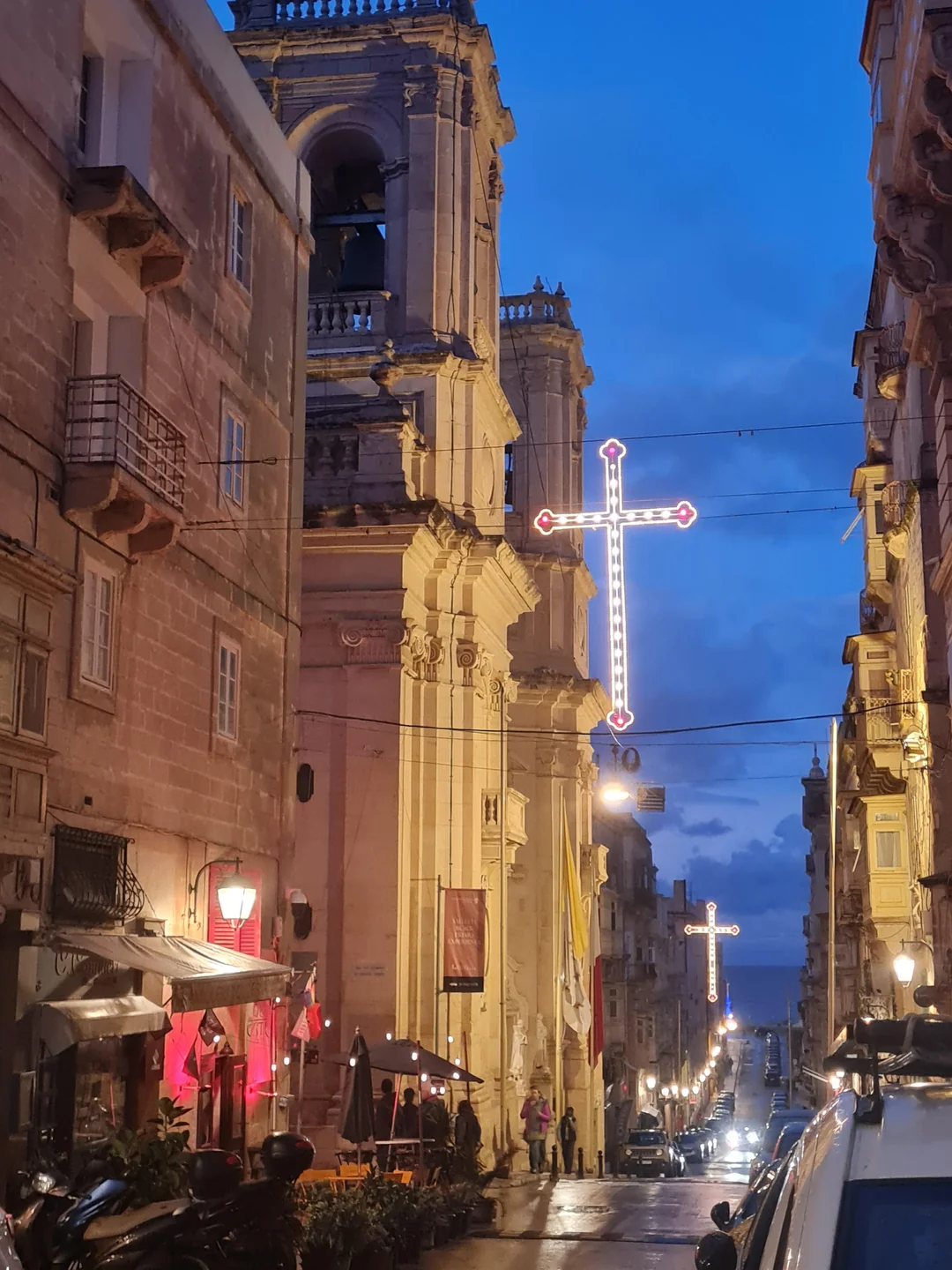 "Evening view of a narrow historic street in Malta at dusk, with a large illuminated cross hanging above the road. Traditional Maltese stone buildings and a grand church tower with a bell tower line the street, lit by warm street lamps. People are gathered near outdoor café tables, and parked cars line the wet road leading toward the sea in the distance."