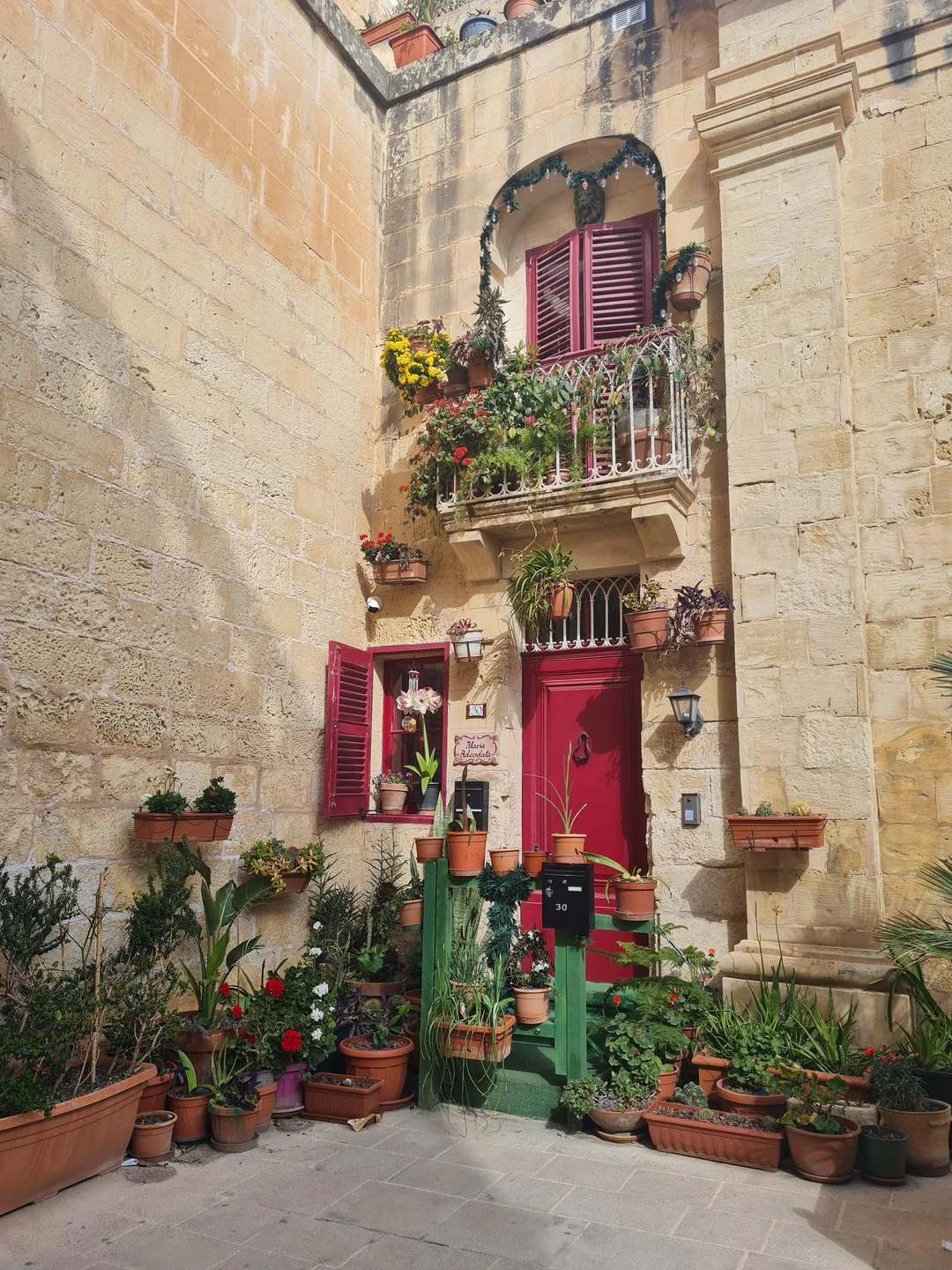 "Colorful courtyard entrance of a Maltese stone house with a bright red door, red shutters, and an abundance of potted flowers and plants on a sunny day."