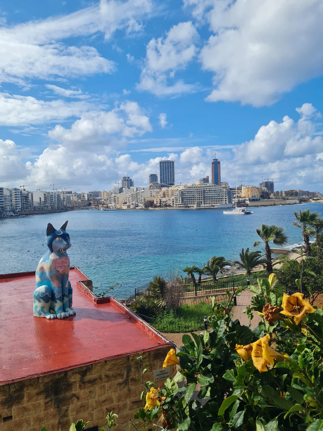 "A colorful cat statue painted in blue and camouflage patterns sits on a bright red rooftop overlooking a stunning panoramic view of Malta’s harbor and modern city skyline. In the background, a white sailboat floats on the turquoise sea under a bright blue sky with scattered clouds. Yellow flowers bloom in the foreground."