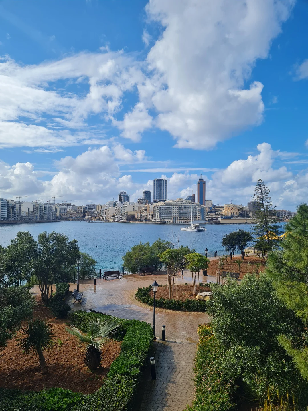 "Beautiful panoramic view over a Maltese harbor with modern city buildings and a sailboat, seen from a green waterfront park under a blue sky."