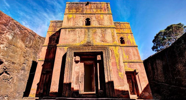 "A low-angle shot of the Church of Saint George in Ethiopia, carved from a single block of pinkish volcanic tuff. The image captures the weathered texture of the stone, narrow windows, and the deep trench surrounding the structure."