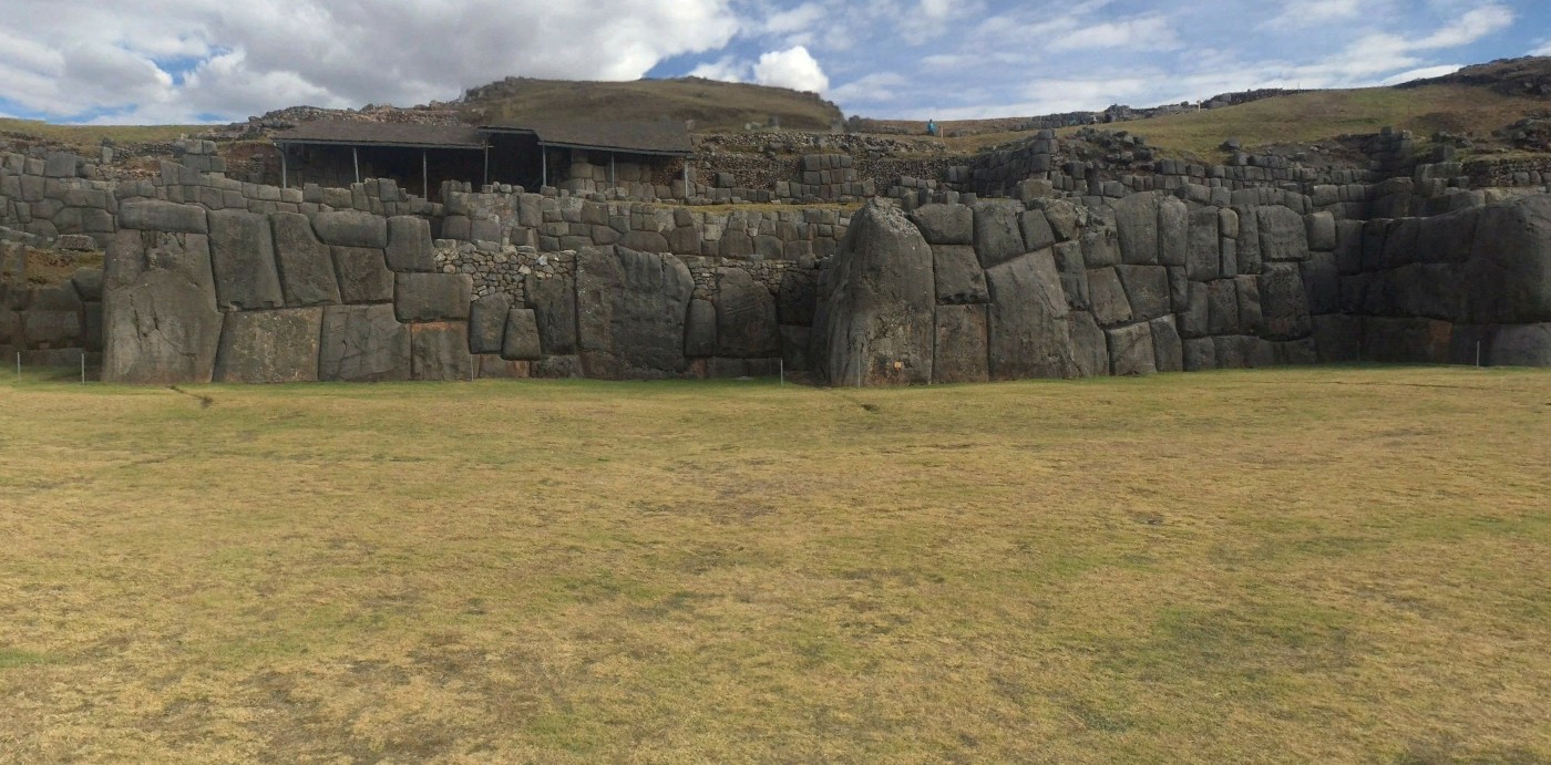 "Massive Inca polygonal stone walls of Sacsayhuamán archaeological site near Cusco, Peru, with a large grassy field in the foreground and a traditional shelter structure on top of the wall under a blue sky with clouds."