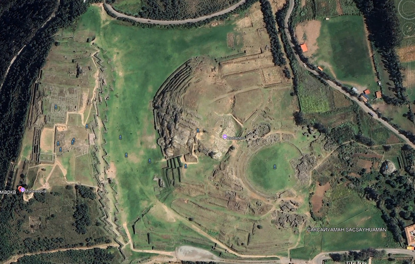 "Aerial satellite view of the ancient Inca archaeological complex of Sacsayhuamán (Sacsayhuaman) near Cusco, Peru, showing the massive zigzag stone walls, large green plazas, terraces, and circular structures."