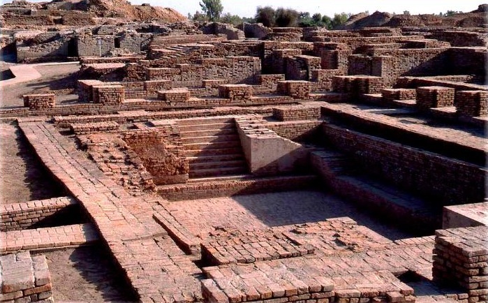 "Ancient ruins of Mohenjo-Daro, showing extensive red brick structures, foundations of houses, narrow streets, and stepped platforms under bright daylight in Pakistan."