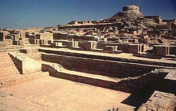 "Extensive ancient stone ruins with tiered terraces, steps, and building foundations in the foreground, leading up a hill to a prominent circular stone tower or citadel under a clear blue sky in an arid landscape."