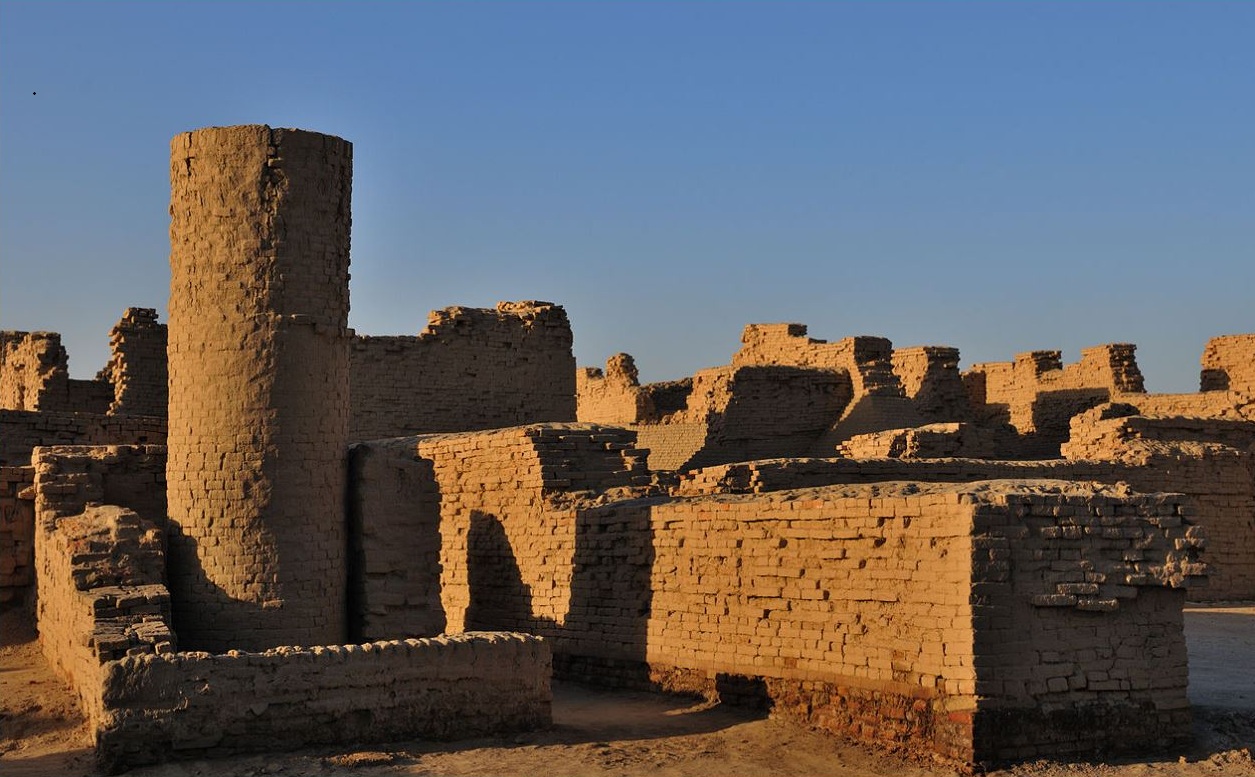"Ancient mud-brick ruins in a desert landscape featuring a tall cylindrical tower and crumbling walls under a clear blue sky."