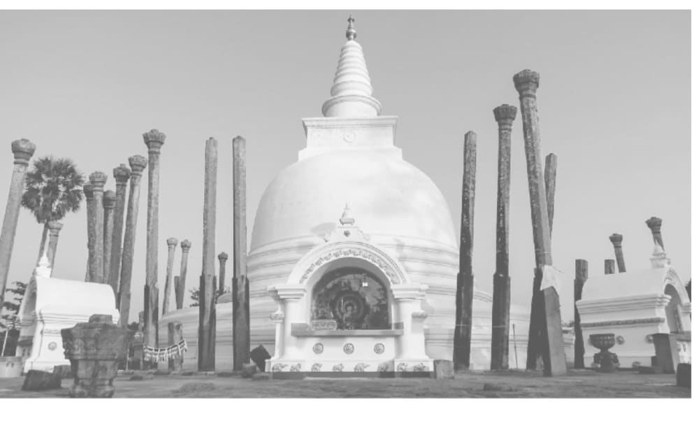 A photograph of a white bell-shaped stupa with a tall spire, surrounded by rows of tall carved stone pillars, some leaning, in an open archaeological site.