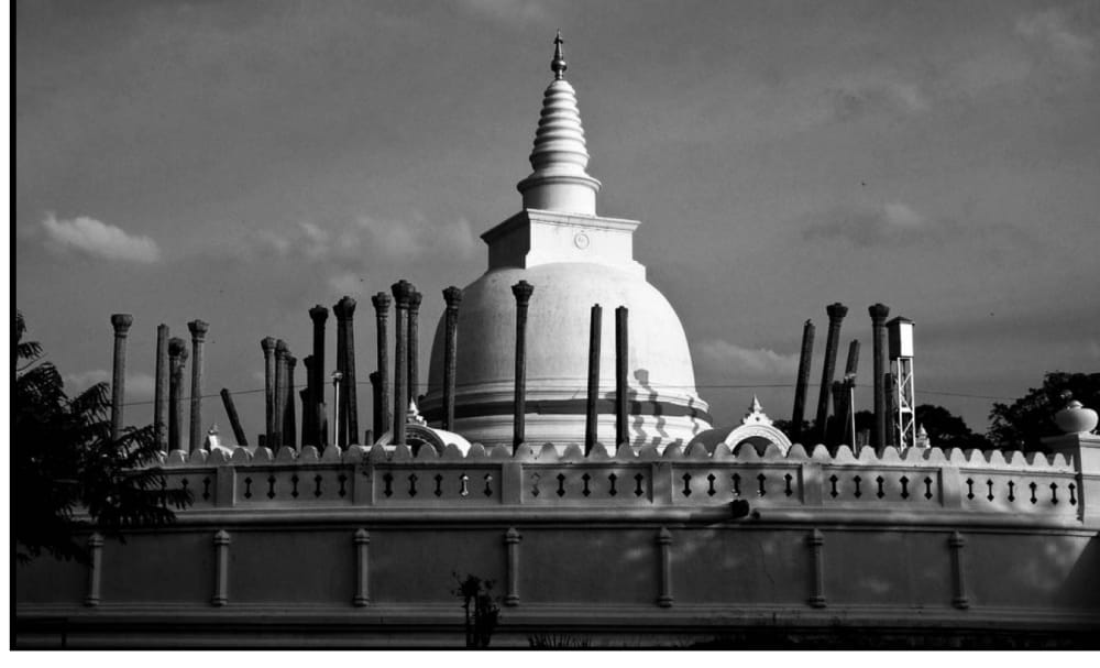 A photograph of a white bell-shaped stupa with a tall spire, seen behind a decorative boundary wall, surrounded by tall stone pillars.