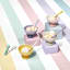 Le Creuset�s Sorbet Collection displayed with a pastel-striped backdrop. The image features colorful packaging for the collection, stacked bowls, mugs, and a footed ice cream bowl with a spoon. The products are arranged on pastel blocks in shades of pink, yellow, and lavender.